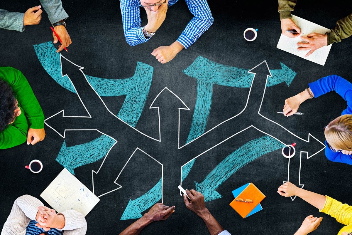 Overhead view of a diverse group of people in a circle around a chalkboard with directional arrows, suggesting collaborative planning.