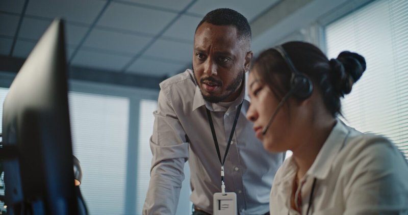 A manager in a button-down shirt leans over a desk to look at a monitor with a colleague wearing a headset in a call center.