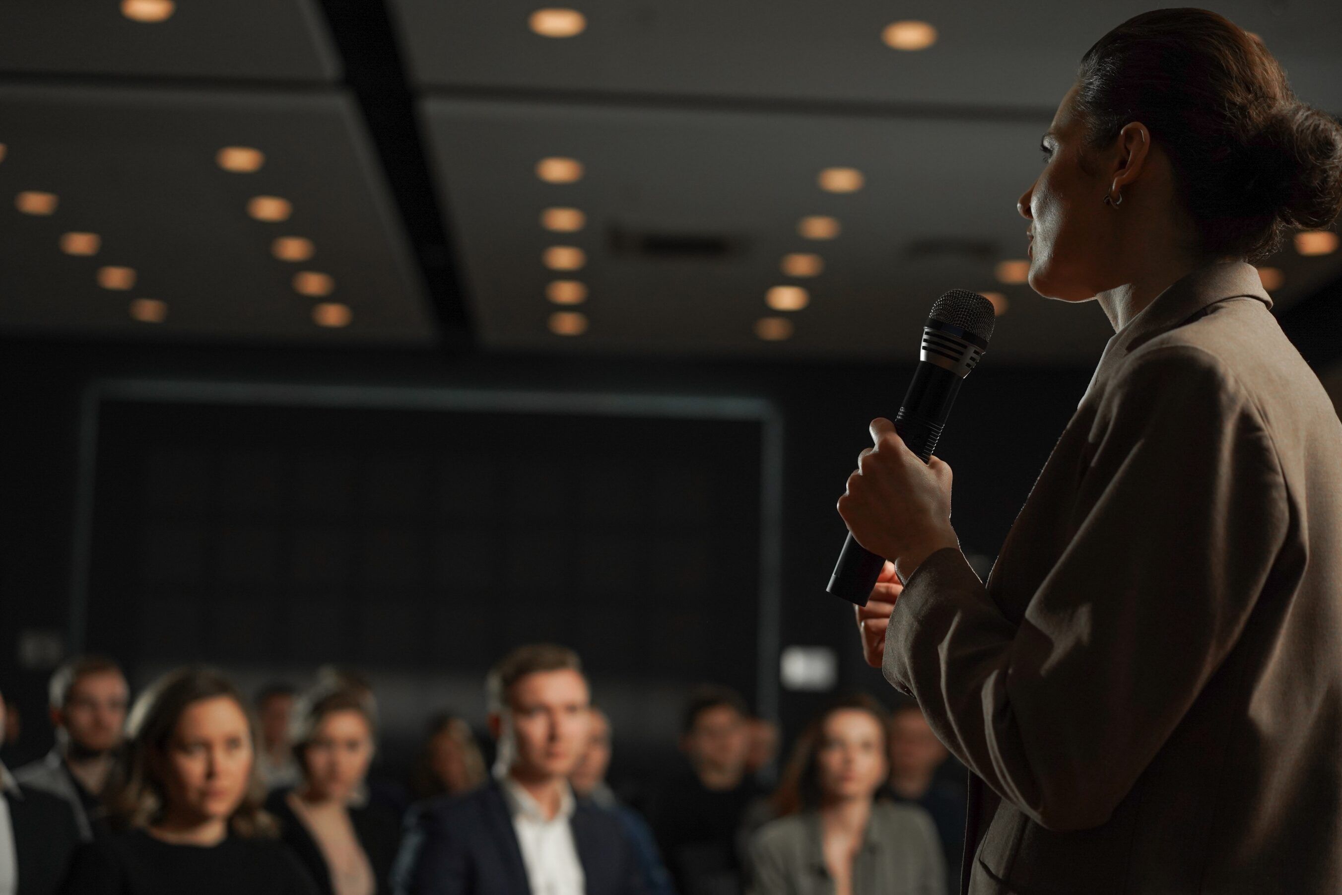 A woman in a tan blazer holds a microphone and addresses an audience in a dimly lit conference hall, with warm ceiling lights overhead and attendees seated in soft focus in the foreground.