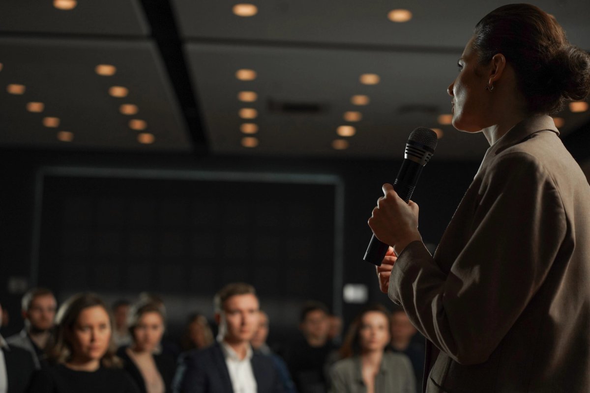 A woman in a tan blazer holds a microphone and addresses an audience in a dimly lit conference hall, with warm ceiling lights overhead and attendees seated in soft focus in the foreground.