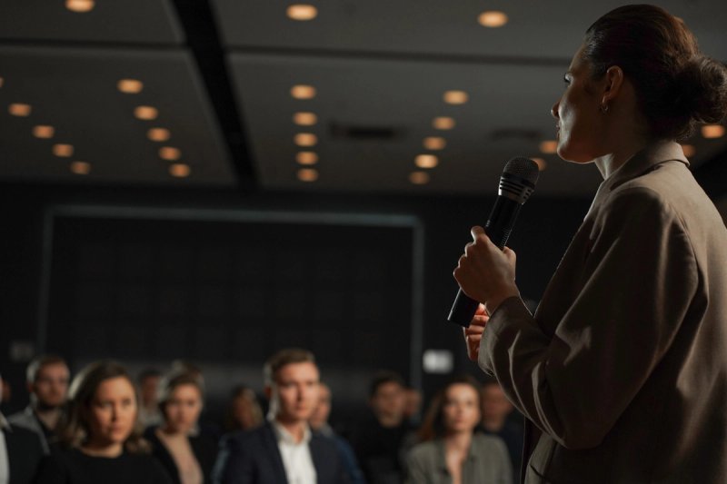 A woman in a tan blazer holds a microphone and addresses an audience in a dimly lit conference hall, with warm ceiling lights overhead and attendees seated in soft focus in the foreground.