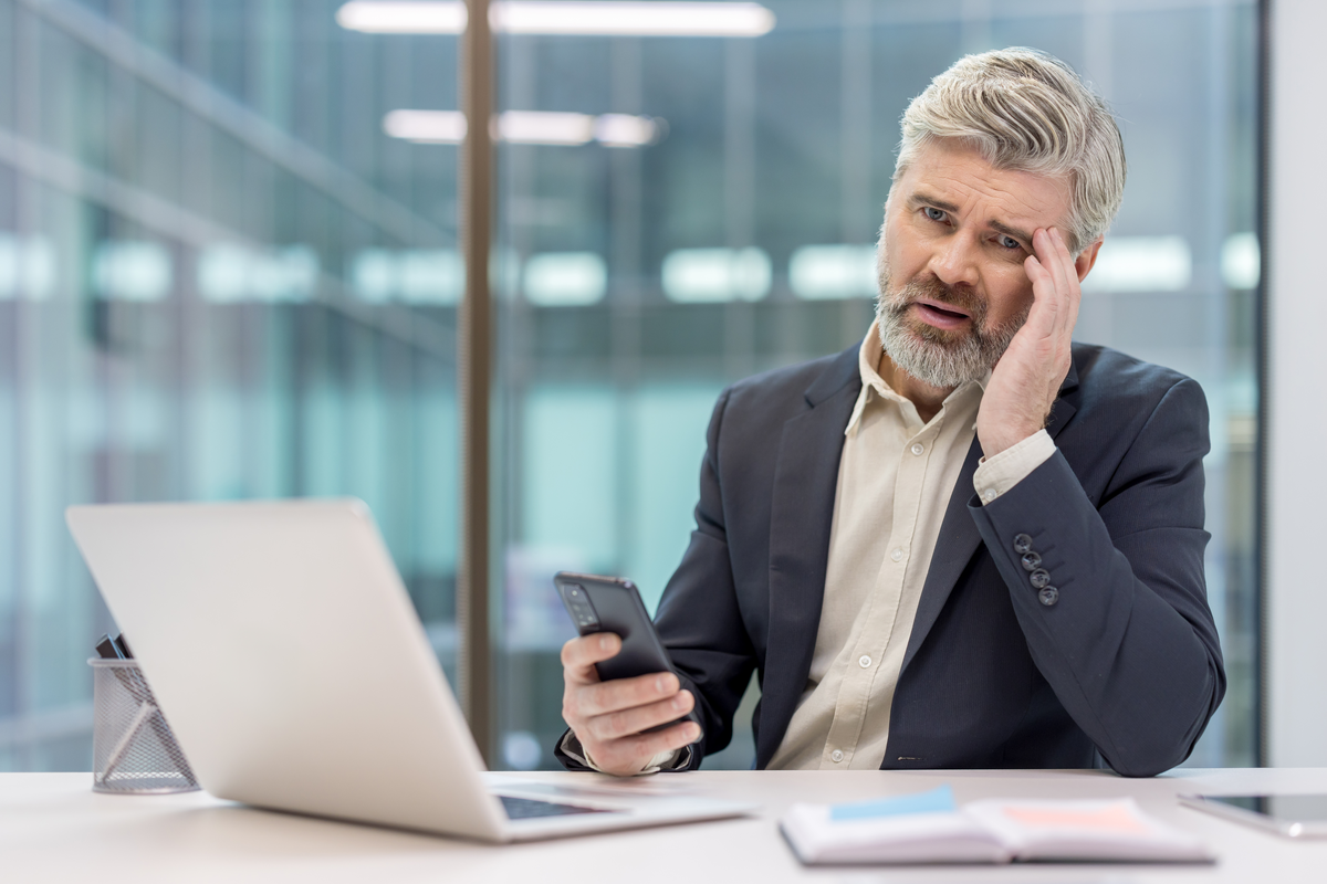 A person with a gray beard sits at a white desk with an open laptop in a glass-walled office, looking at a phone.