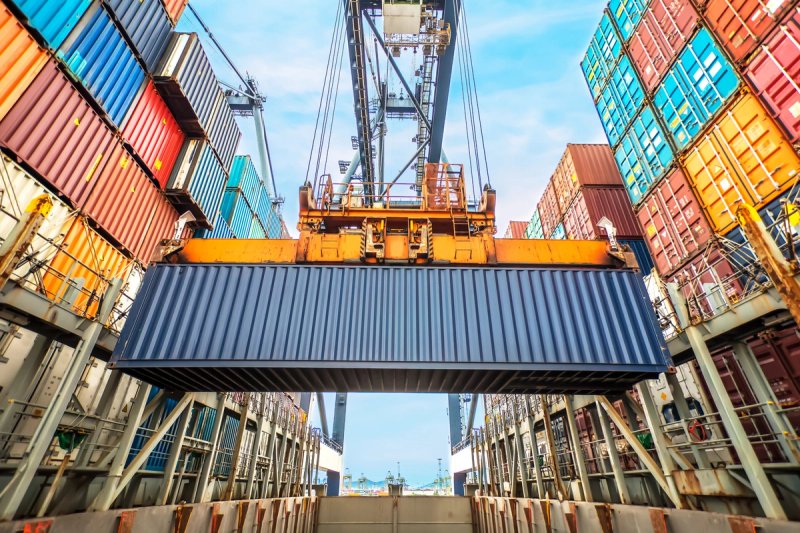 A crane lifts a large blue shipping container above stacked rows of colorful containers at a busy port under a partly cloudy sky.