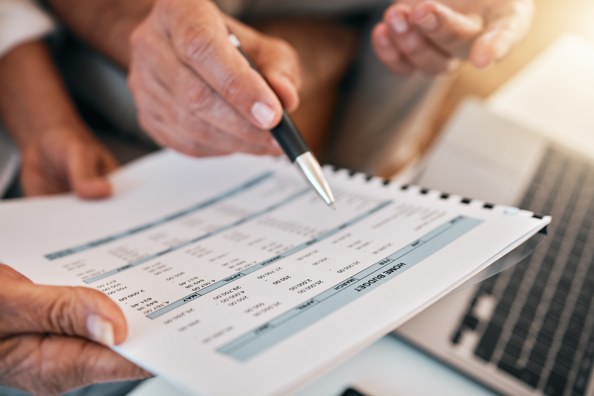 Close-up of a hand using a pen to point at a row in a printed financial spreadsheet held by another person, with an open laptop visible on the desk beside them.