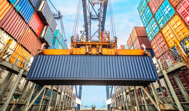 Yellow crane lifting a dark blue cargo container at a busy international shipping port terminal.