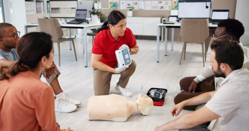 A trainer in a red shirt kneels on the floor and demonstrates an automated external defibrillator next to a CPR mannequin while seated learners watch.