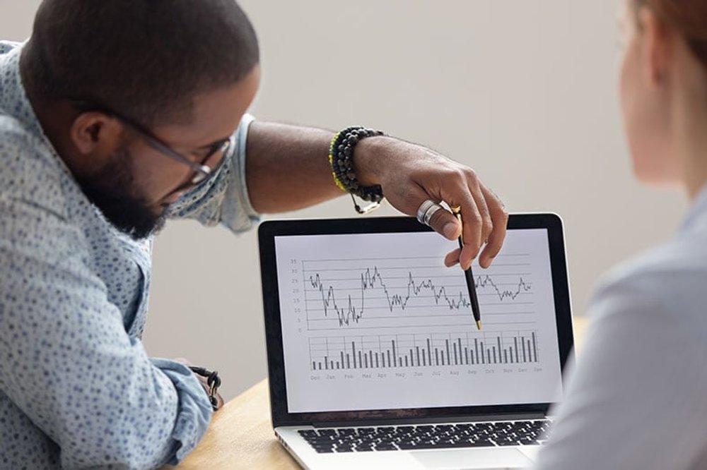Man points with pen at a bar chart on a laptop, while coworker watches.