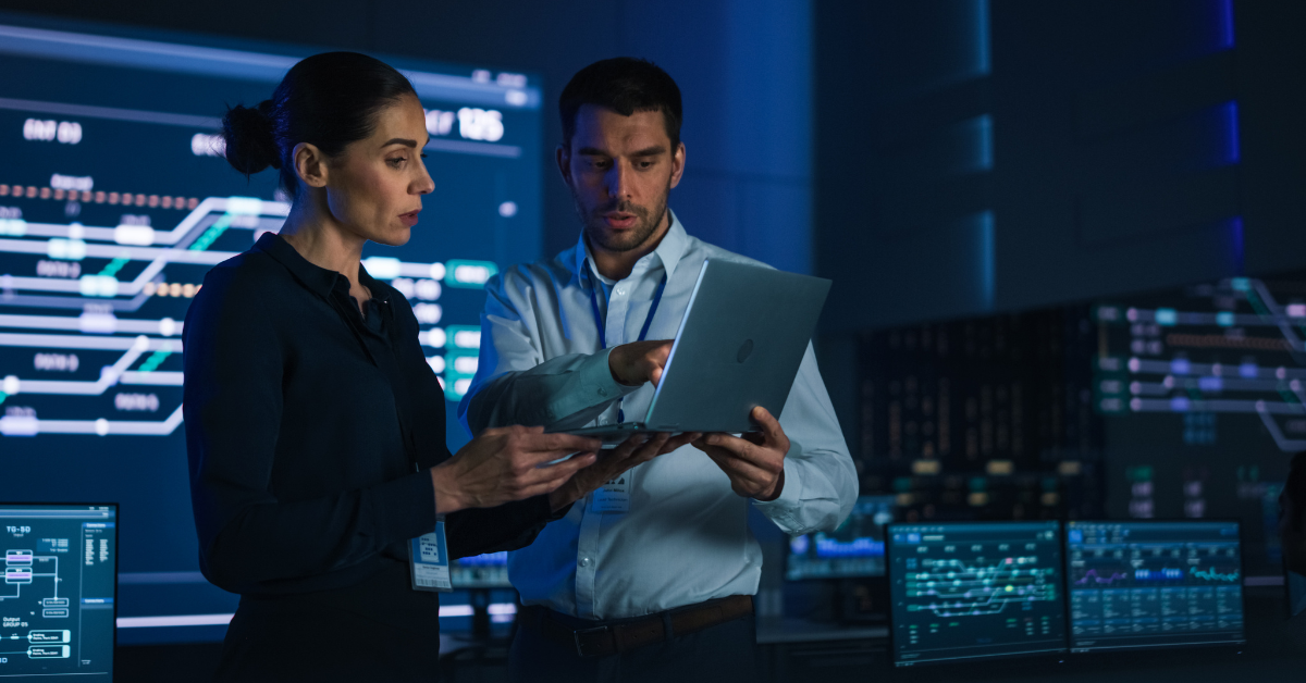 Two professionals reviewing a tablet in a high-tech control room with blue digital monitoring displays in the background.