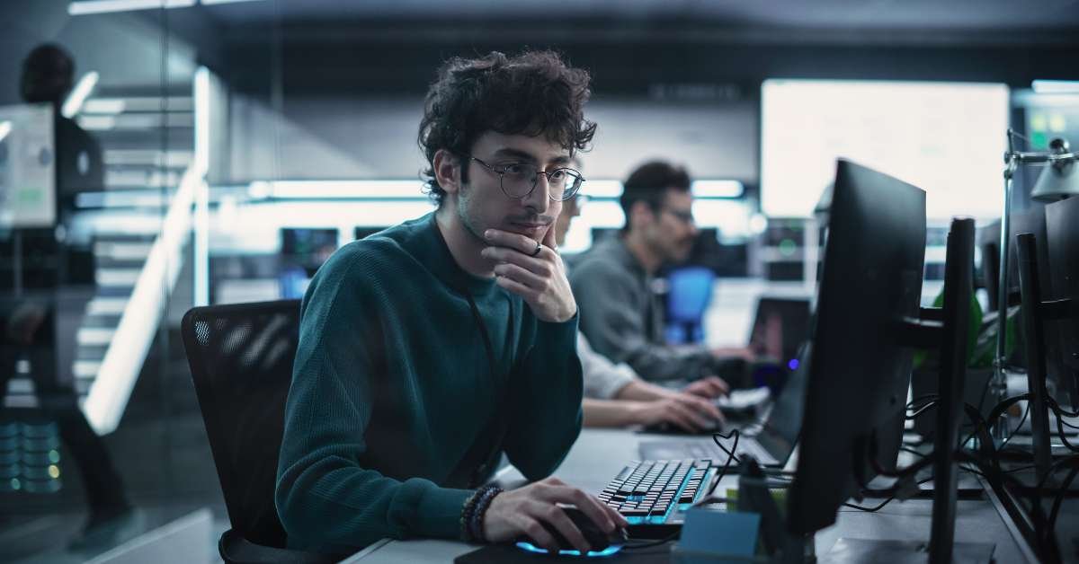 Young man with glasses working at a computer in a busy open-plan tech office with multiple monitors and neon lighting.