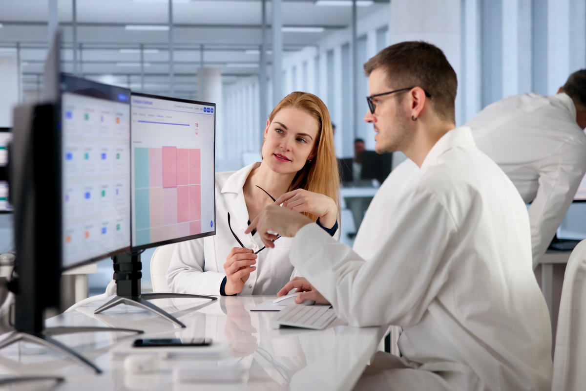 Two people in white lab coats sit at a workstation reviewing charts on large monitors in a bright, modern open-plan office.