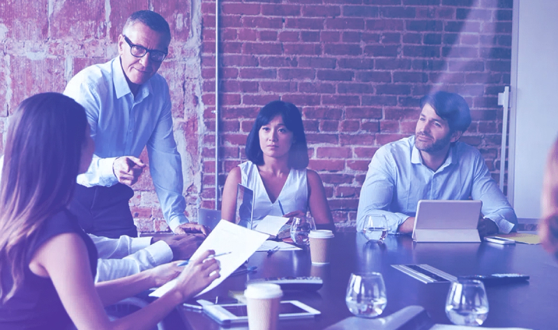 Four professionals collaborating around a conference table in front of an exposed brick wall, with laptops and documents on the table.