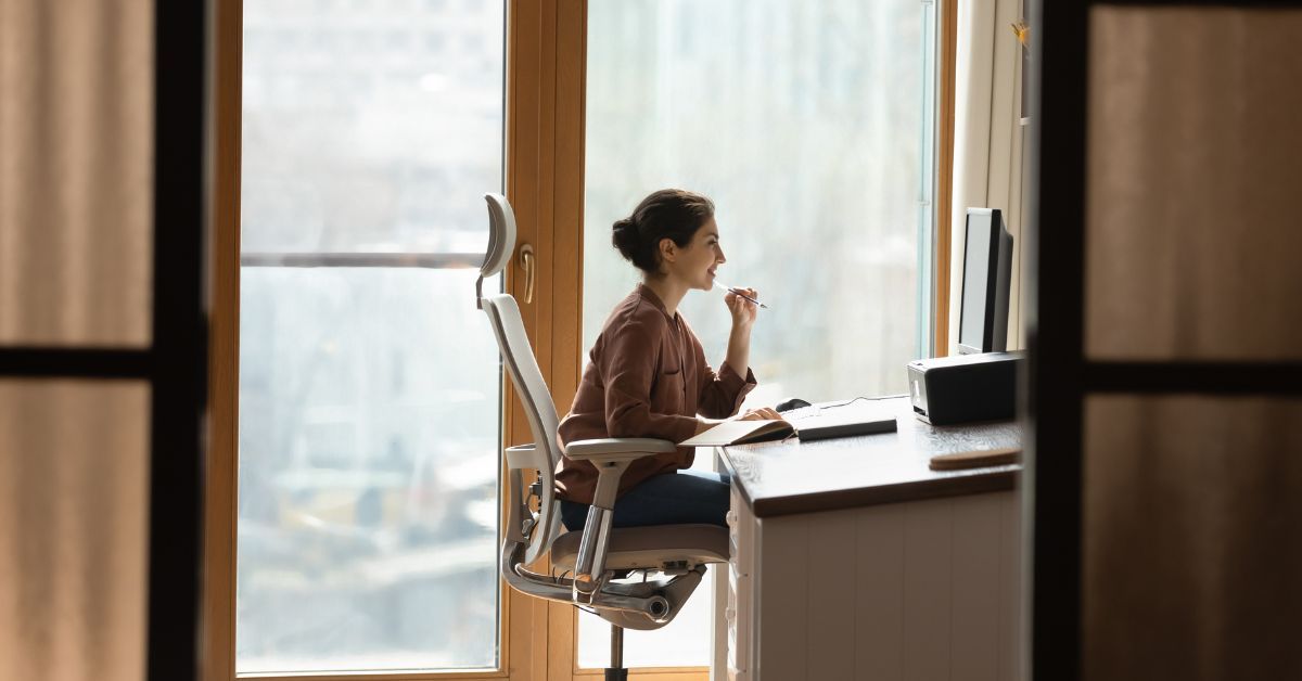 Woman in a burgundy shirt working at a desk in a high-rise office with floor-to-ceiling windows and a city view.