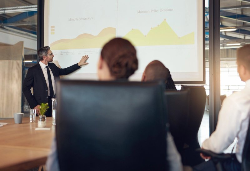 A presenter in a black suit gestures toward a projected line chart while colleagues watch from a conference table.