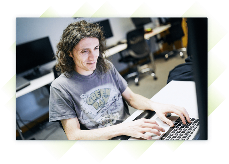 A team member wearing a t-shirt, smiling while working on a laptop at a desk.