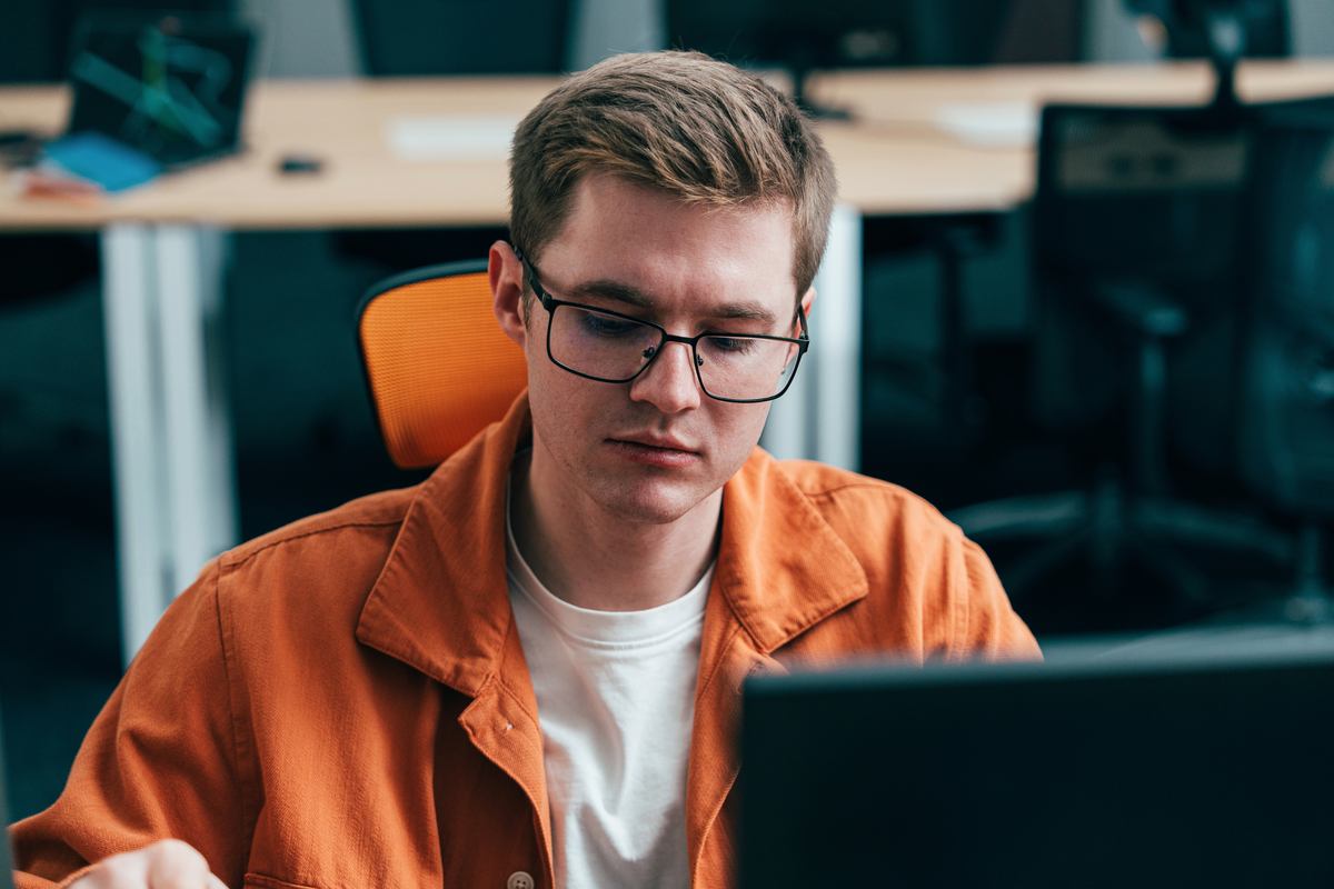 A person in an orange button-up shirt over a white t-shirt and black-framed glasses works at a laptop in an open-plan office, with empty desks and chairs visible in the background.