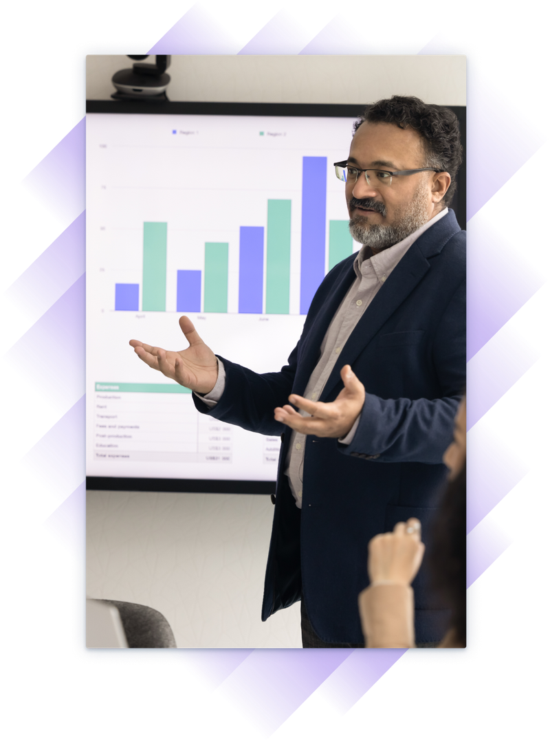 Executive discussing financials with team in boardroom, charts visible on screen behind him as he gestures.