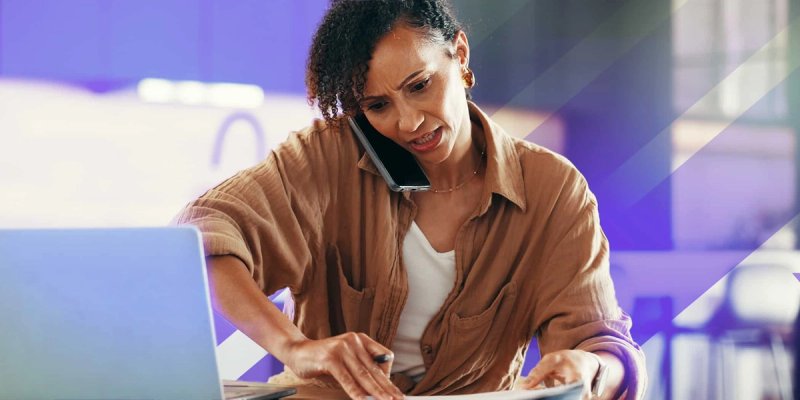 A training professional multitasks at a desk, holding a phone between her ear and shoulder while working on paperwork and using a laptop. She looks focused and slightly stressed because she is using an LMS to wrangle ILT.