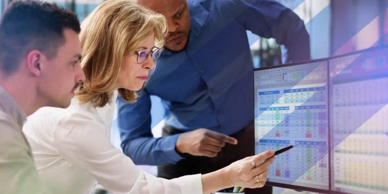 Three professionals closely reviewing complex data on a dual-monitor setup, highlighting collaboration and data-driven decision-making—ideal for illustrating enterprise training management or system comparison.