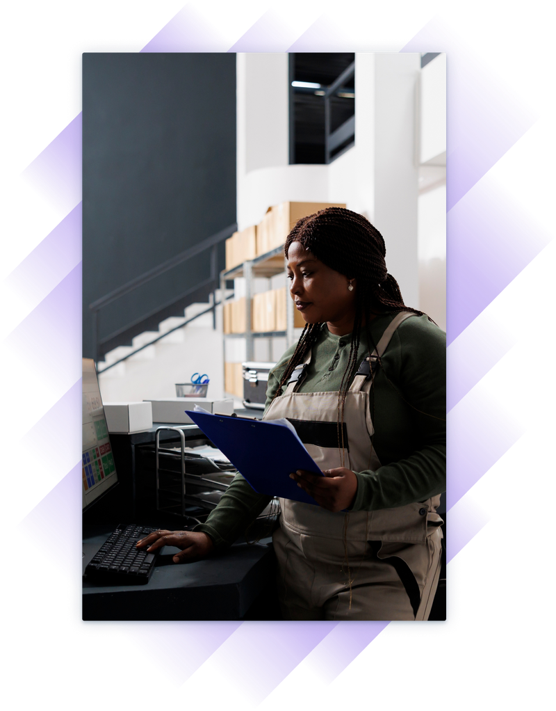 Woman in work overalls using a desktop computer while holding a clipboard in a stockroom.