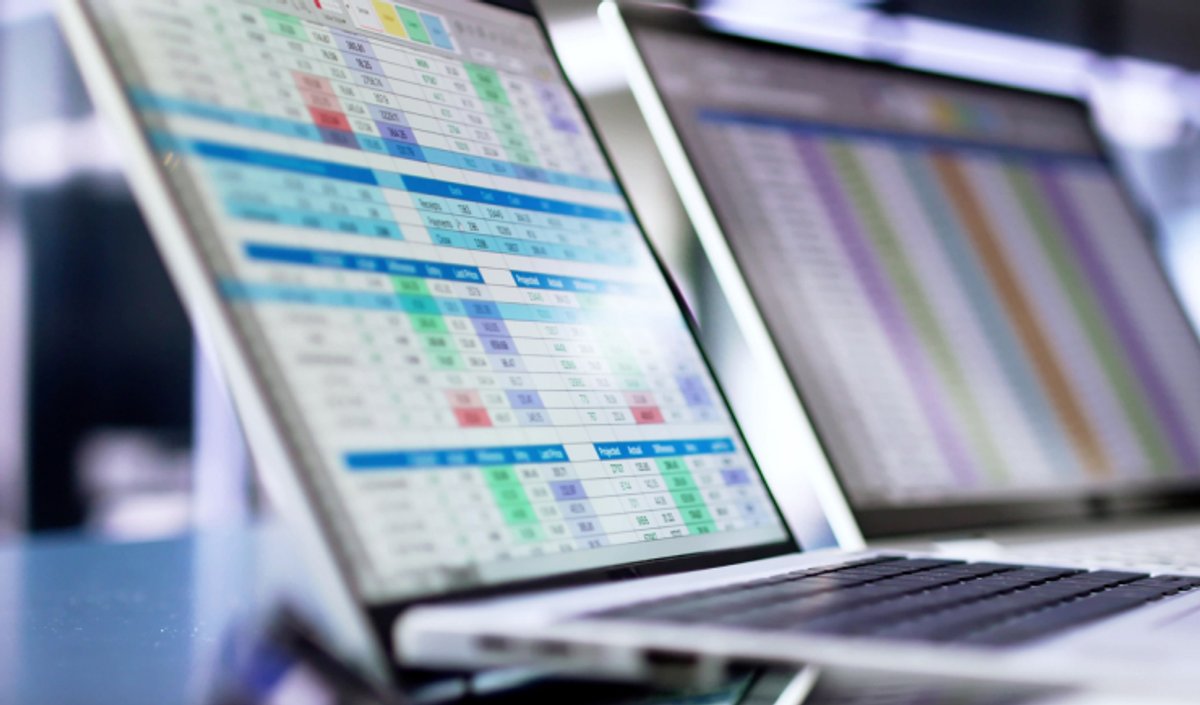 Two laptops side-by-side in an office showing data tables of company financials.
