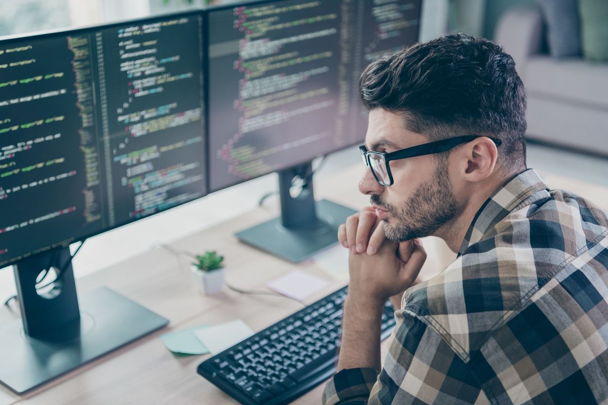 A person in a plaid flannel shirt and glasses rests their chin on their hand while reading code displayed across two large monitors at a bright home workstation.