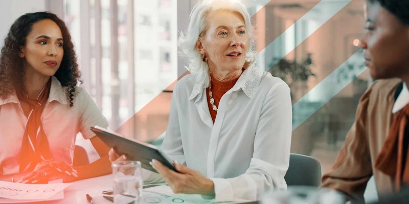 Three professionals sit around a conference table having a discussion. One woman holds a tablet while the others listen attentively to her describing the benefits of a TMS and LMS together.