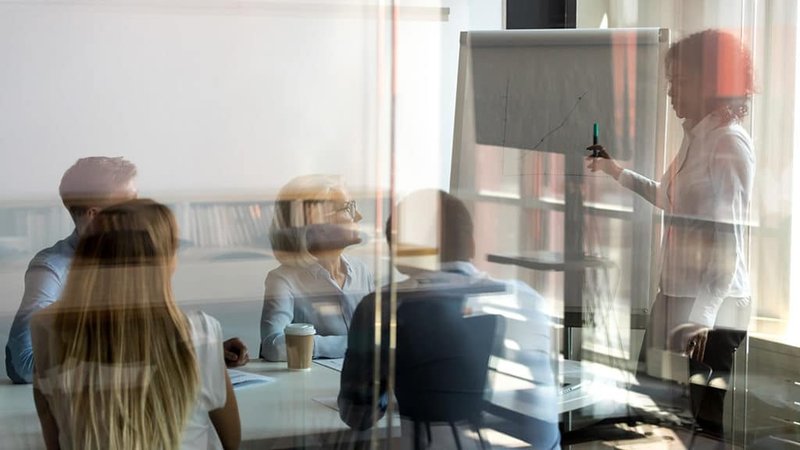 Woman pointing at a chart drawn onto a large pad while presenting findings to a room full of her colleagues.