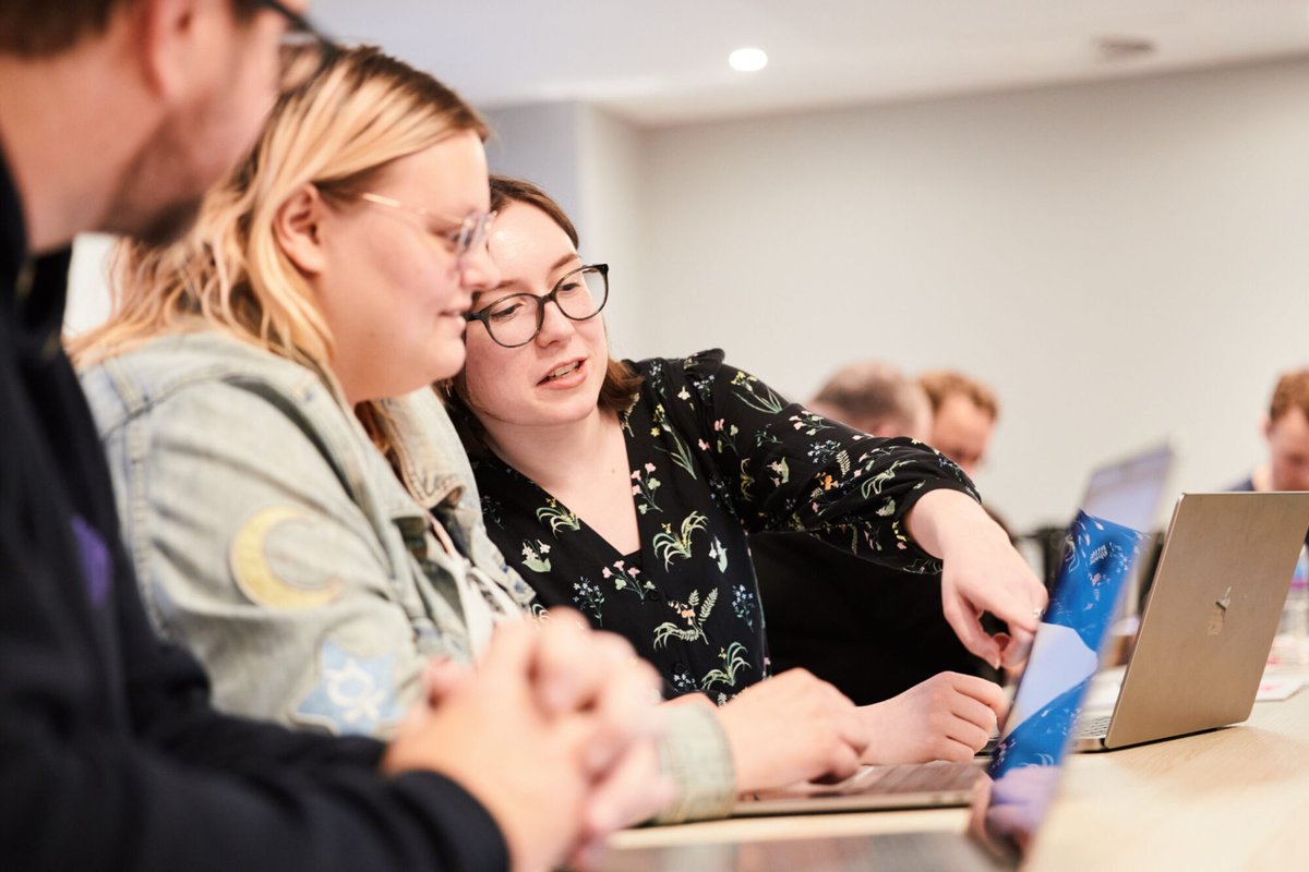 Two Administrate software engineers sitting around a laptop while one instructs the other by pointing at the screen.