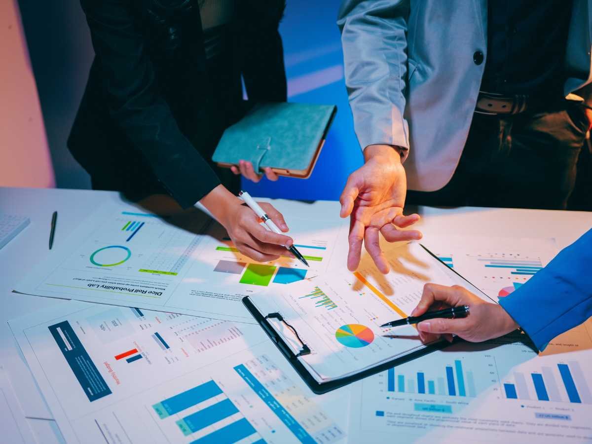 Three people's hands point at and highlight various colorful bar charts, pie graphs, and data reports spread across a desk, illustrating a collaborative team review of performance or compliance metrics.
