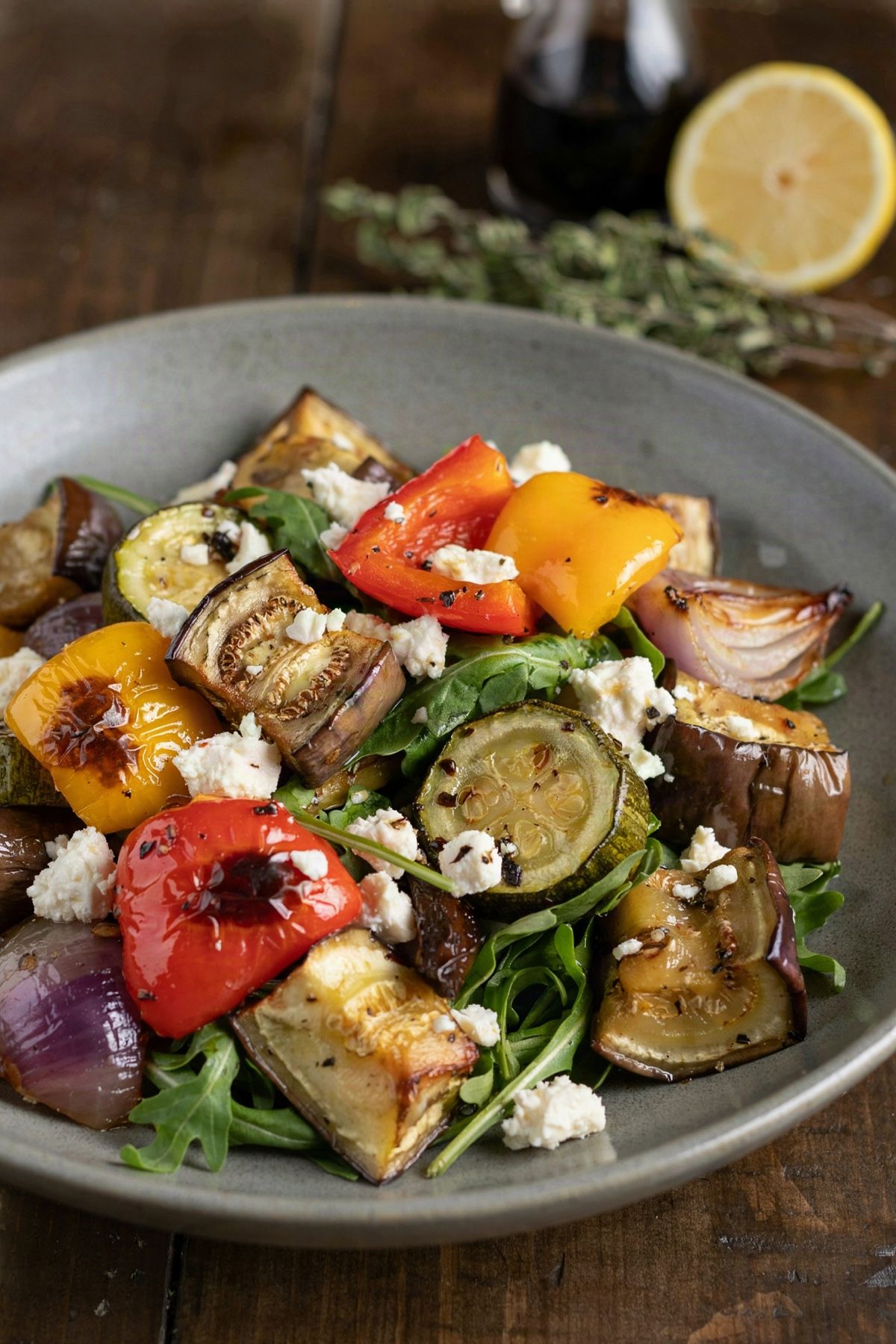 Mediterranean roasted vegetable salad in a grey stoneware bowl: browned chunks of eggplant, red and yellow pepper, zucchini and red onion tossed with arugula, glossy with balsamic and scattered with crumbled feta