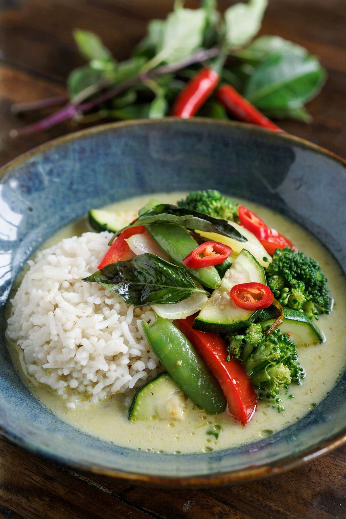 Vegan Thai green curry in a slate blue bowl, broccoli, red pepper, zucchini and snap peas in a pale green coconut sauce beside jasmine rice, topped with Thai basil and sliced red chili