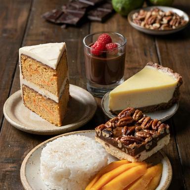Five vegetarian desserts on a dark wooden table: a slice of two-layer carrot cake under off-white frosting, a glass of dark chocolate mousse topped with raspberries, a wedge of pale yellow key lime pie on a dark crumb crust, a pecan pie bar, and mango with sticky rice.
