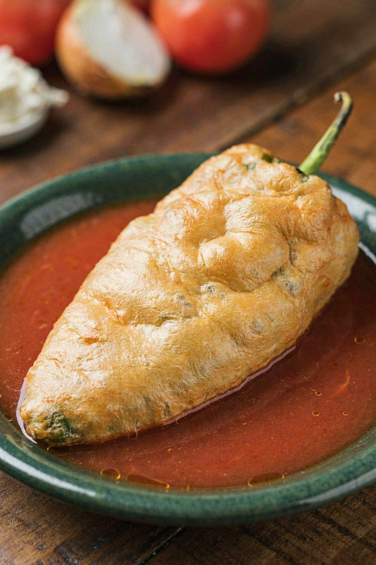 A Mexican chile relleno in a shallow green bowl, a whole poblano in puffed golden egg batter sitting in thin red tomato caldillo.