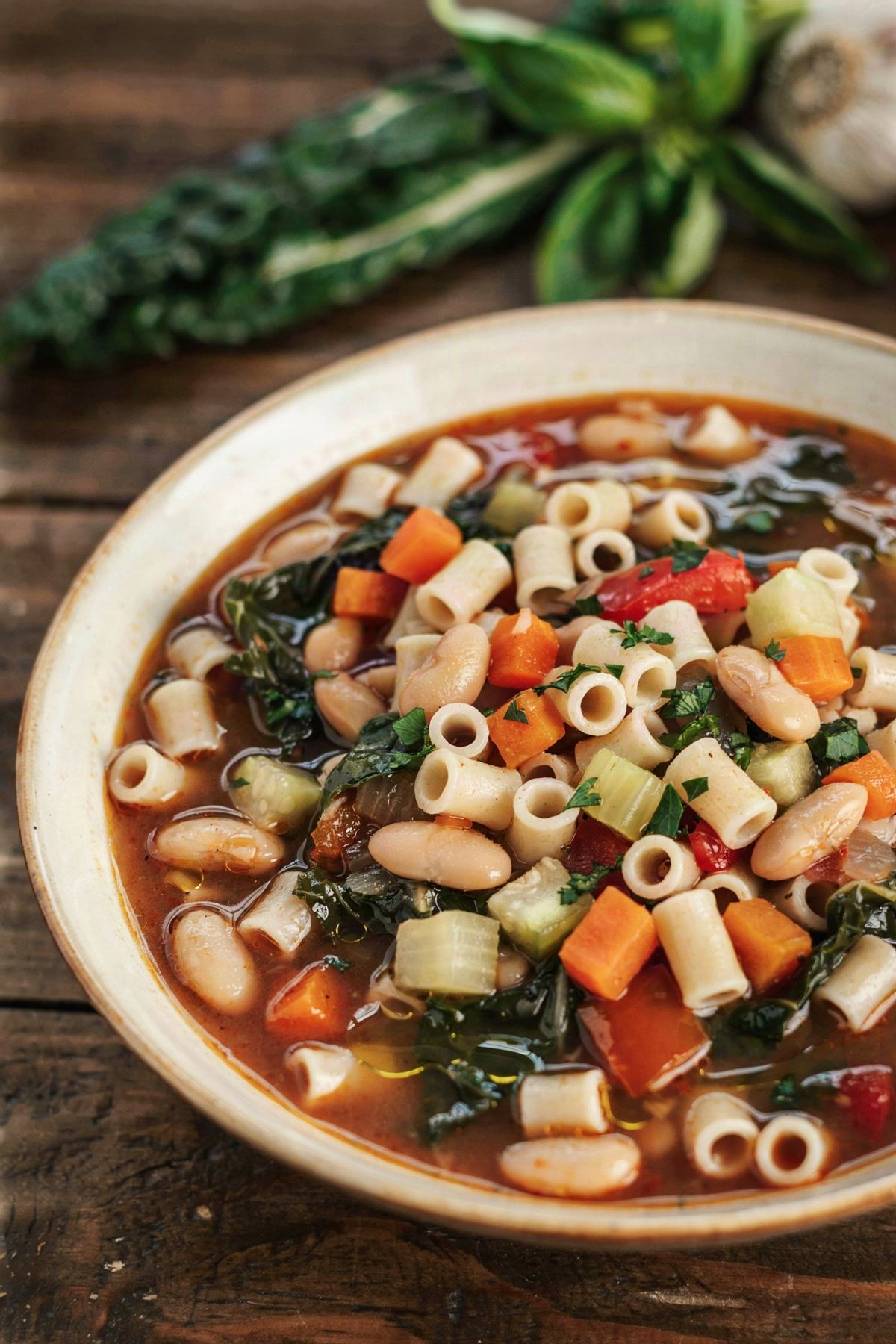 Vegan minestrone soup in a cream bowl, a brick red broth with cannellini beans, ditalini pasta, diced carrot, celery and zucchini, red bell pepper and dark green kale, finished with a drizzle of olive oil and chopped parsley.