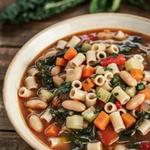 Vegan minestrone soup in a cream bowl, a brick red broth with cannellini beans, ditalini pasta, diced carrot, celery and zucchini, red bell pepper and dark green kale, finished with a drizzle of olive oil and chopped parsley.