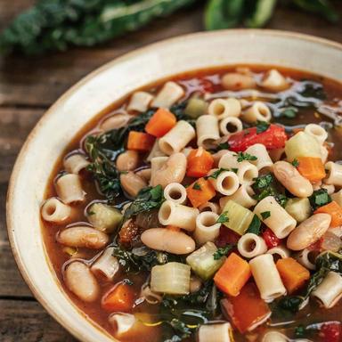 Vegan minestrone soup in a cream bowl, a brick red broth with cannellini beans, ditalini pasta, diced carrot, celery and zucchini, red bell pepper and dark green kale, finished with a drizzle of olive oil and chopped parsley.