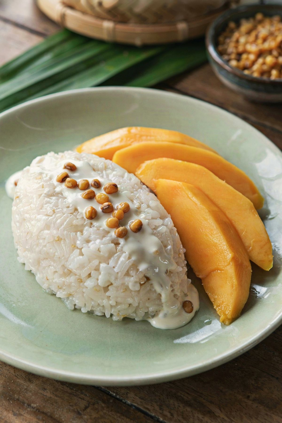Thai mango sticky rice on a celadon plate, a mound of glossy coconut sticky rice under poured salted coconut cream with toasted mung beans, beside fanned slices of ripe golden mango.