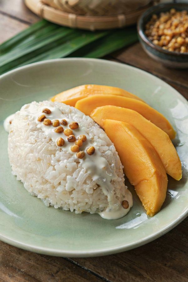 Thai mango sticky rice on a celadon plate, a mound of glossy coconut sticky rice under poured salted coconut cream with toasted mung beans, beside fanned slices of ripe golden mango.