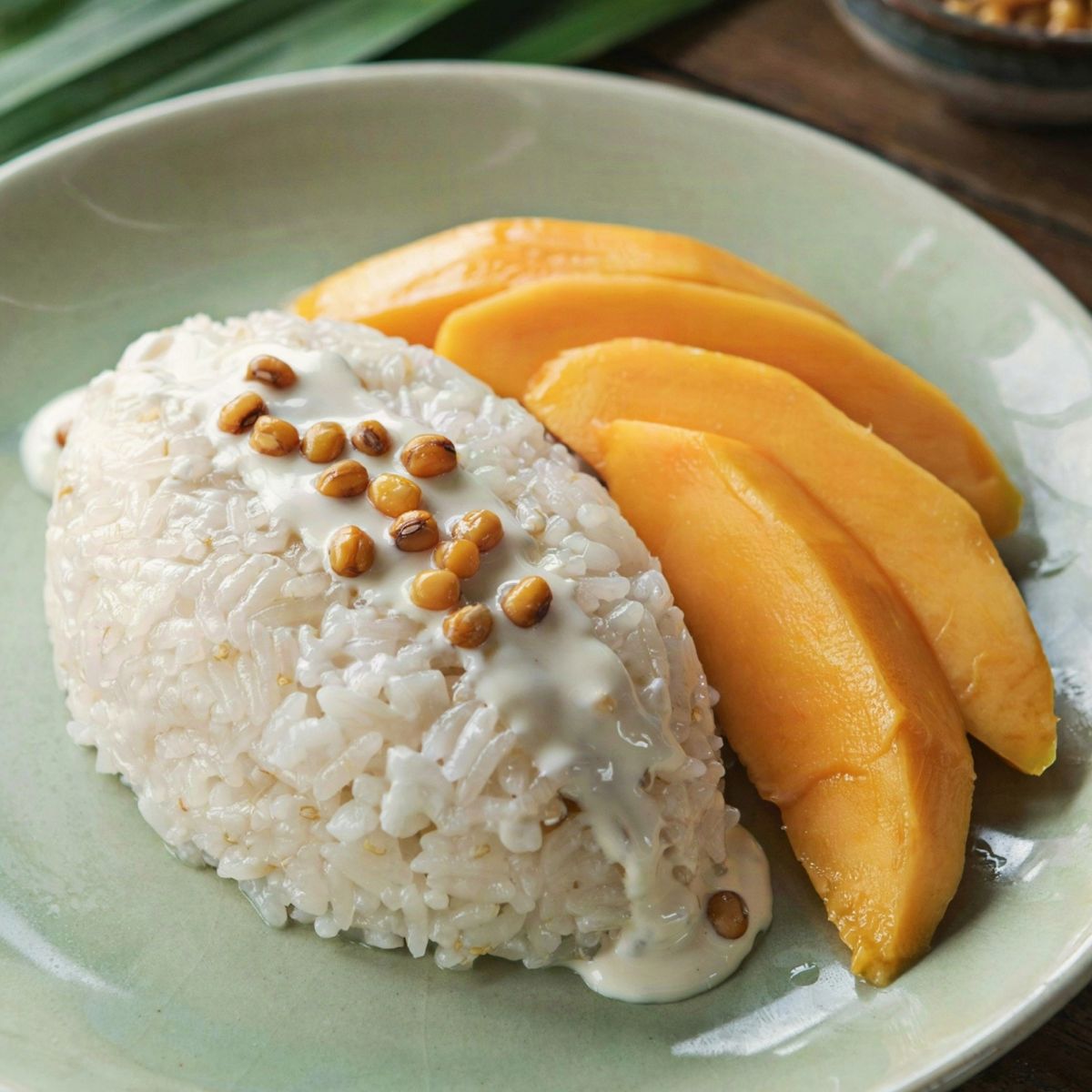 Thai mango sticky rice on a celadon plate, a mound of glossy coconut sticky rice under poured salted coconut cream with toasted mung beans, beside fanned slices of ripe golden mango.