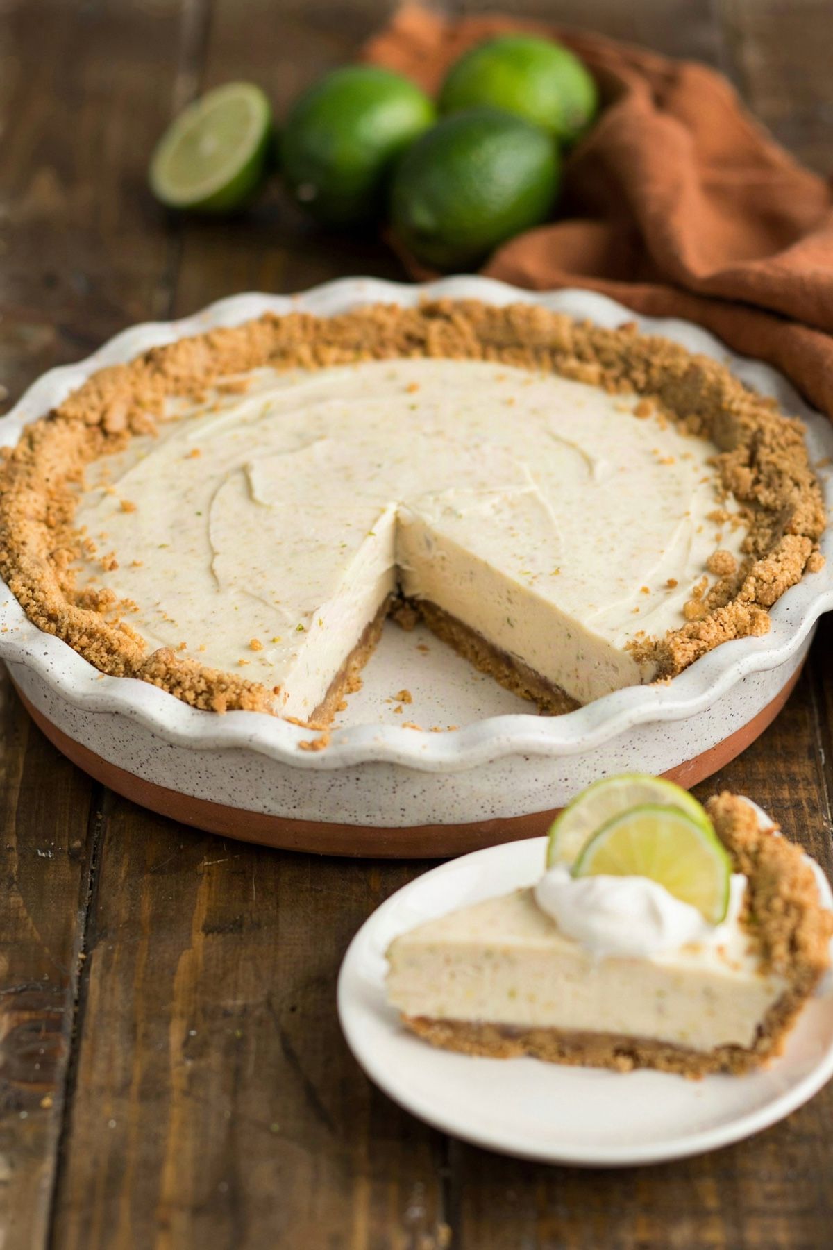 A whole vegan key lime pie in a speckled ceramic dish with one slice cut away, the pale ivory filling set firm on a golden graham cracker crust, the slice plated with coconut whipped cream and lime rounds.