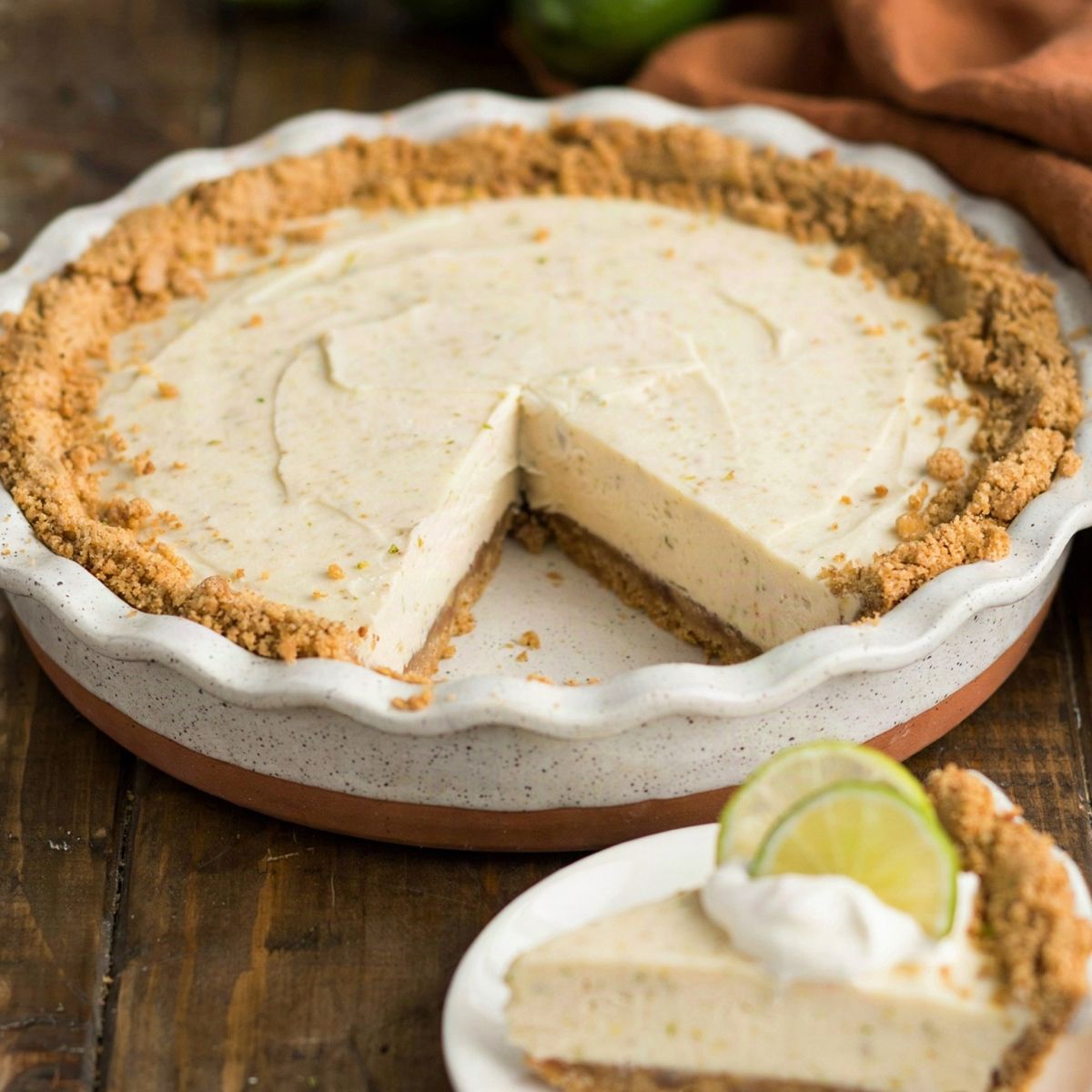 A whole vegan key lime pie in a speckled ceramic dish with one slice cut away, the pale ivory filling set firm on a golden graham cracker crust, the slice plated with coconut whipped cream and lime rounds.