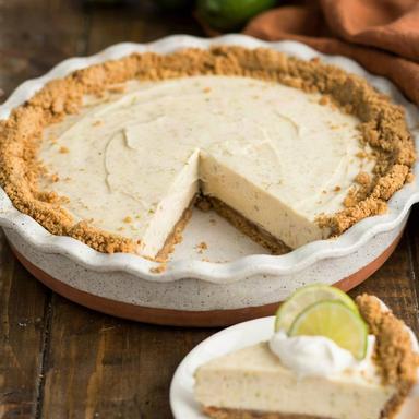 A whole vegan key lime pie in a speckled ceramic dish with one slice cut away, the pale ivory filling set firm on a golden graham cracker crust, the slice plated with coconut whipped cream and lime rounds.