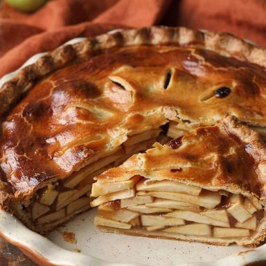 Vegan apple pie in a white speckled ceramic dish with one wedge cut away, the deep golden crust opened to show layers of pale apple slices set in a glossy amber filling
