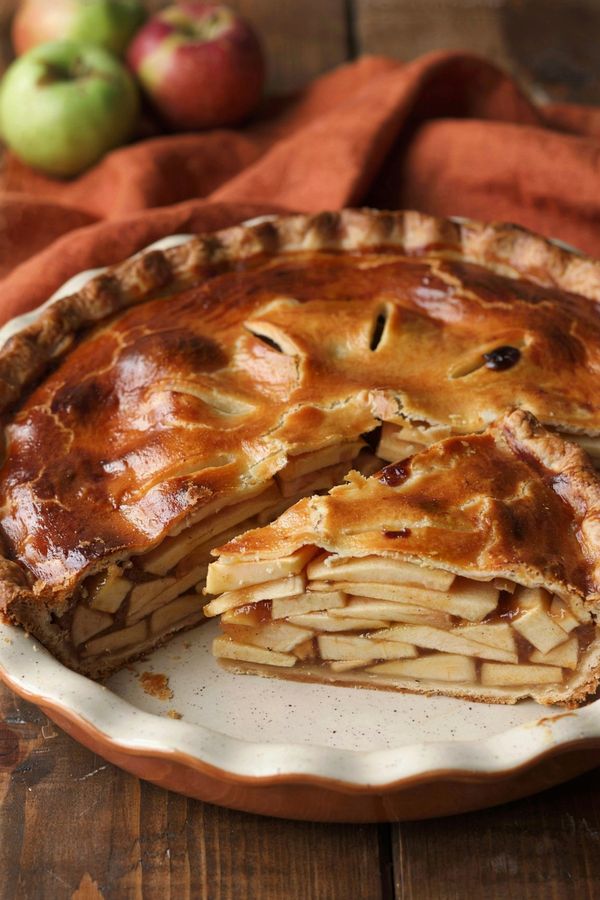 Vegan apple pie in a white speckled ceramic dish with one wedge cut away, the deep golden crust opened to show layers of pale apple slices set in a glossy amber filling