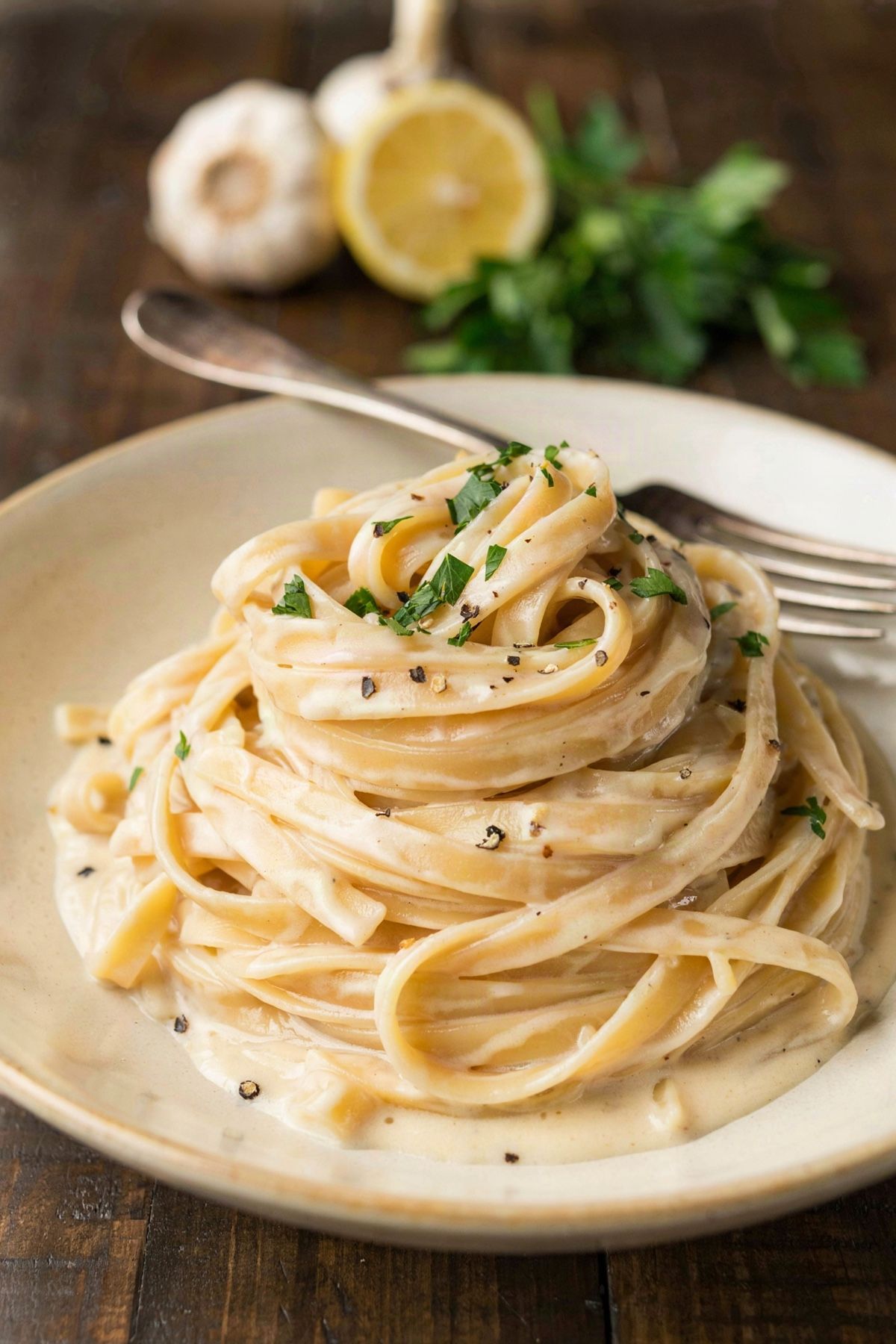 Vegan fettuccine Alfredo in a cream stoneware bowl, long ribbons coated in a glossy pale cashew cream sauce with chopped parsley and cracked black pepper, garlic and lemon behind