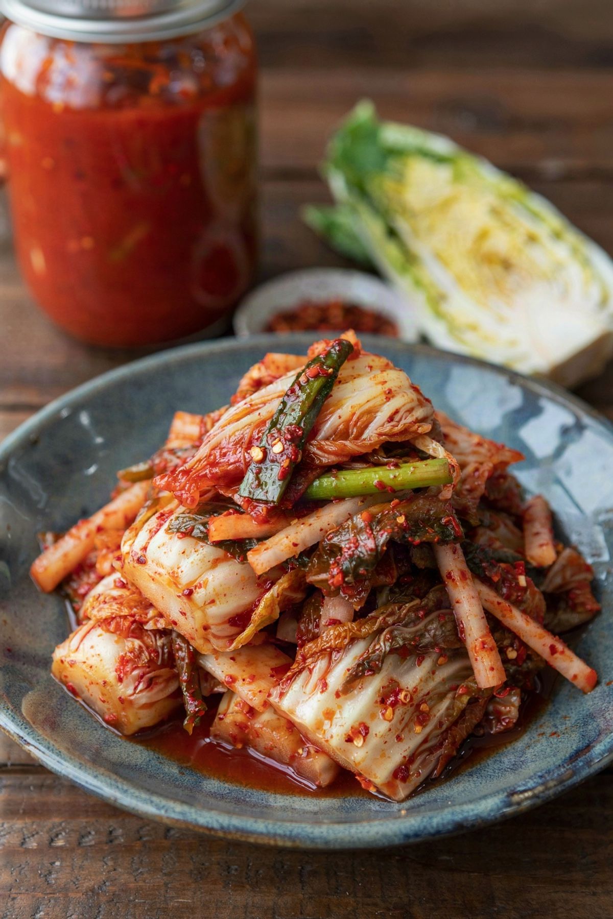 Vegetarian kimchi heaped in a dark blue-gray ceramic bowl, pieces of napa cabbage coated in coarse brick-red gochugaru paste with radish matchsticks and scallion lengths, a glass jar of kimchi and half a napa cabbage behind.