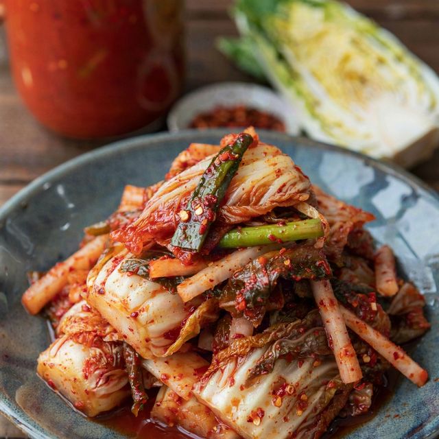 Vegetarian kimchi heaped in a dark blue-gray ceramic bowl, pieces of napa cabbage coated in coarse brick-red gochugaru paste with radish matchsticks and scallion lengths, a glass jar of kimchi and half a napa cabbage behind.
