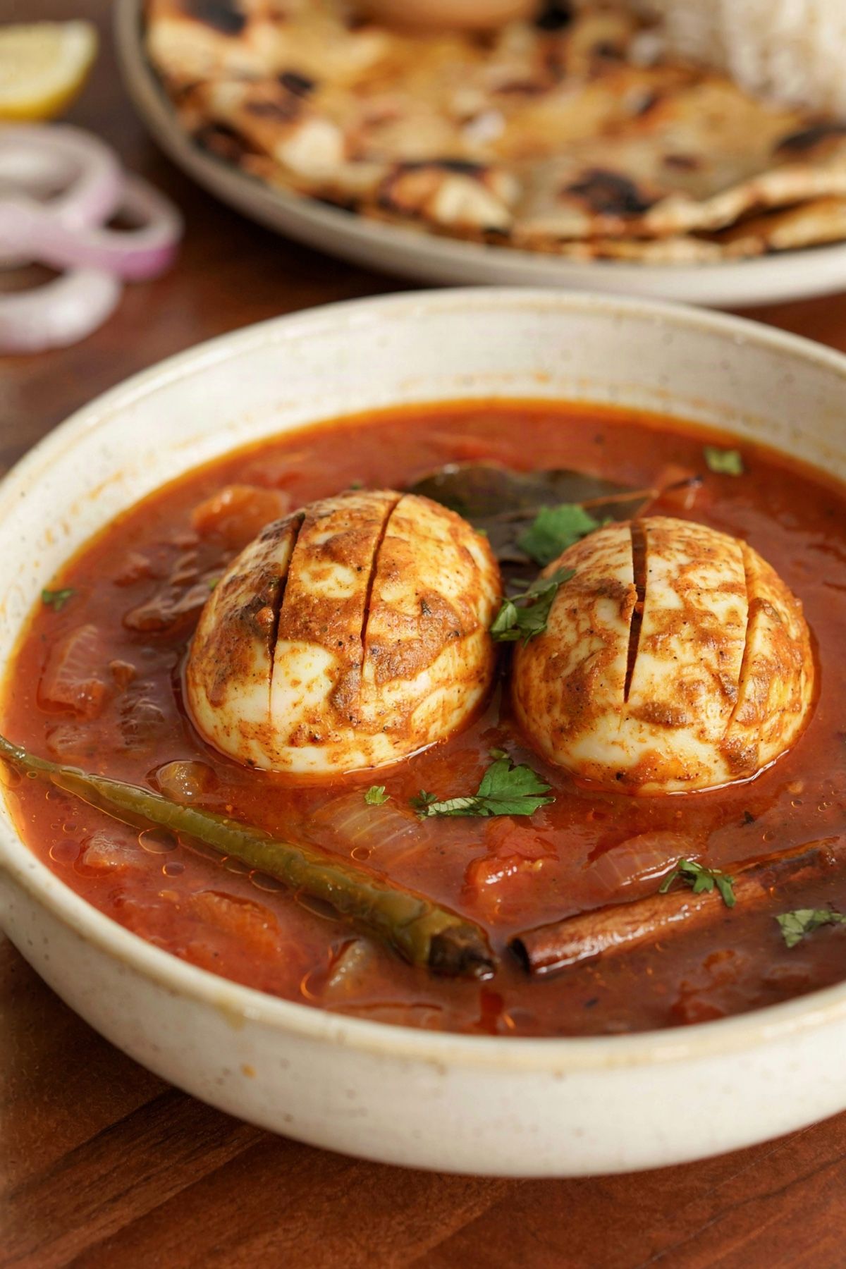 Indian egg curry in a cream ceramic bowl, two whole hard-boiled eggs scored and fried until golden, sitting in a thick brick red onion and tomato gravy with cilantro, green chile and a cinnamon stick, served with roti and rice.