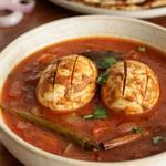 Indian egg curry in a cream ceramic bowl, two whole hard-boiled eggs scored and fried until golden, sitting in a thick brick red onion and tomato gravy with cilantro, green chile and a cinnamon stick, served with roti and rice.