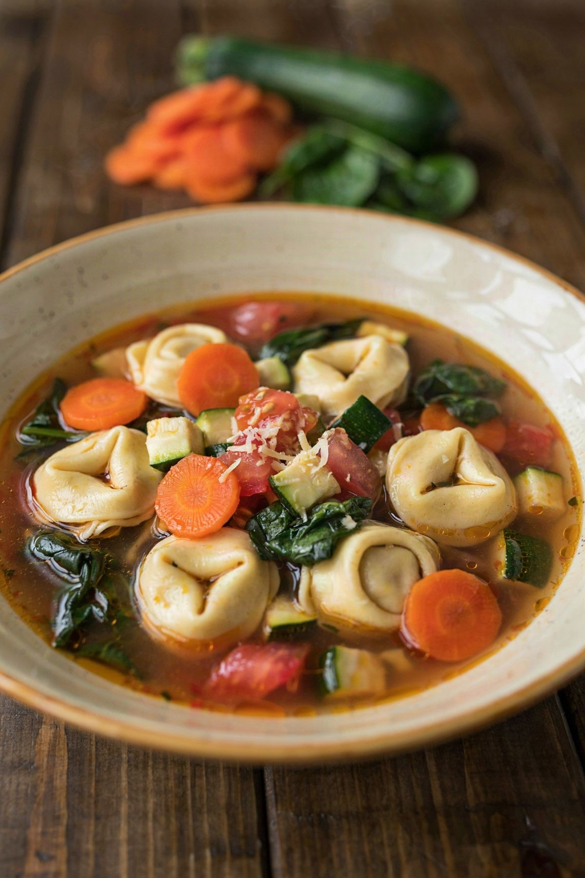 A bowl of meatless tortellini soup: cheese tortellini, carrot discs, zucchini and spinach in a clear tomato broth, topped with grated cheese.